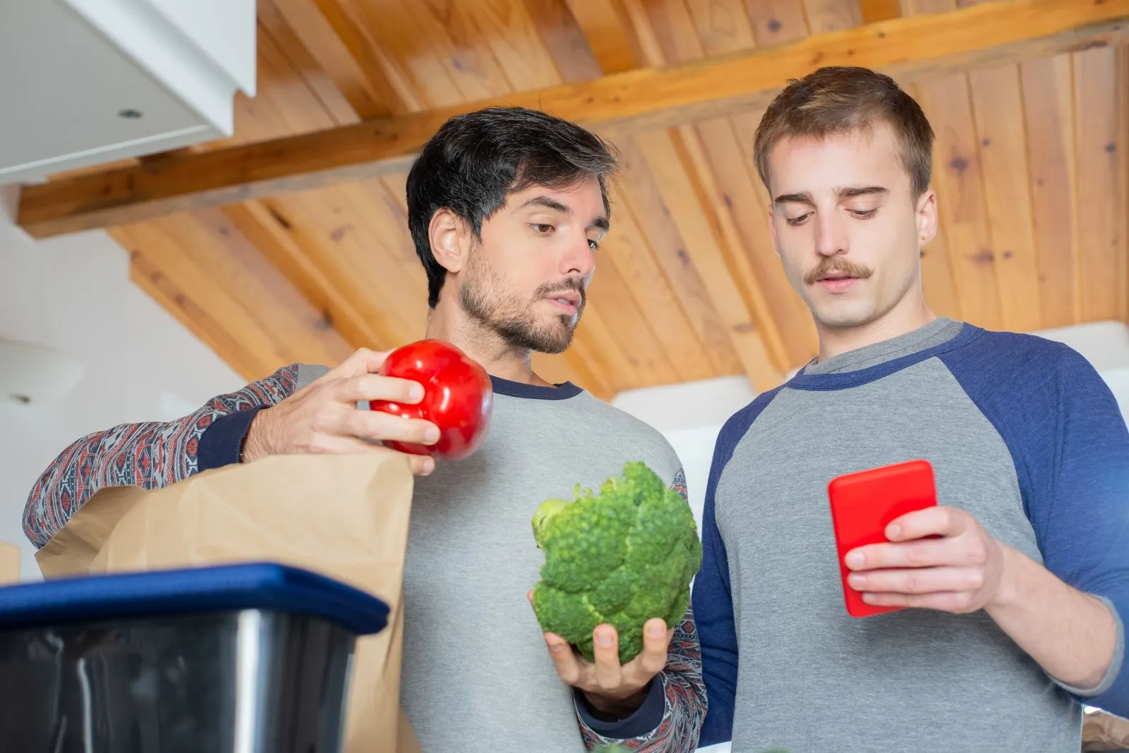 Two men organizing groceries in a kitchen while using a smartphone.